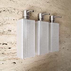 Könnte beinhalten: Drei transparente, strukturierte Seifenspender mit silberfarbenen Pumpköpfen sind an einer hellen, strukturierten Wand befestigt. Die Spender sind rechteckig mit vertikalen Rillen. Für den Einsatz im Badezimmer.