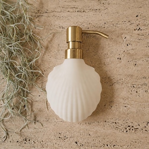 May include: A white soap dispenser shaped like a seashell with a metallic gold pump mechanism. The body of the dispenser features a ribbed, fan-like texture resembling a scallop shell, providing a distinct visual profile for bathroom or kitchen use.