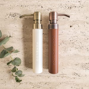 Può includere: Due dispenser ricaricabili per shampoo e lozione per il corpo. Il flacone dello shampoo è bianco con una pompa dorata, e il flacone della lozione per il corpo è marrone con una pompa di rame. Le parole "SHAMPOO" e "BODY LOTION" sono stampate verticalmente.