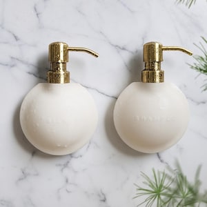 Könnte beinhalten: Zwei kugelförmige weiße Seifenspender mit metallisch goldenen Pumpen. Der linke Spender ist mit dem Wort SILK und der rechte Spender mit dem Wort SHAMPOO geprägt. Beide Behälter haben eine glatte Oberfläche mit sichtbaren Wassertropfen.