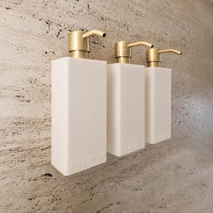 Op de afbeelding: Drie rechthoekige witte zeepdispensers met goudkleurige pompmechanismen. Elke fles heeft in reliëf aangebrachte tekst op de voorzijde, specifiek gelabeld SHAMPOO, SILK en BODY, gerangschikt in een rij tegen een gestructureerde beige stenen muur.