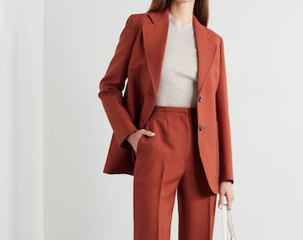 May include: A rust-colored suit featuring a blazer with two buttons and wide-leg trousers. The model is wearing a light-colored top and white sandals. A white handbag with a gold-colored clasp is held in the hand.