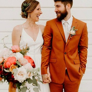 Könnte beinhalten: Ein Paar lächelt sich am Hochzeitstag an. Die Braut trägt ein weißes, ärmelloses Kleid und der Bräutigam trägt einen orangebraunen Anzug.