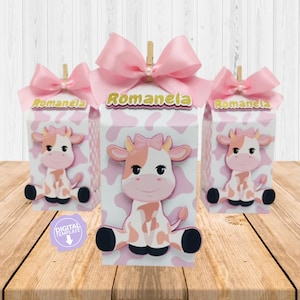 May include: Three rectangular favor boxes featuring a white and pink cow print pattern. Each box is topped with a light pink satin bow and a wooden clip. The front of each box displays a cartoon cow illustration and the name Romanela in gold.
