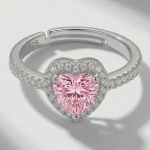 May include: A silver-toned adjustable ring featuring a central heart-shaped pink gemstone surrounded by a halo of small clear stones. The band is stamped with 925 and includes additional clear stones along the sides of the ring shank.