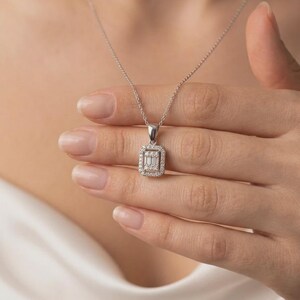 Peut inclure: Un collier argenté doté d'un pendentif rectangulaire avec une pierre centrale taille baguette entourée d'un halo de petites pierres rondes. Le pendentif est suspendu à une chaîne délicate, présentant un design classique aux éclats clairs.