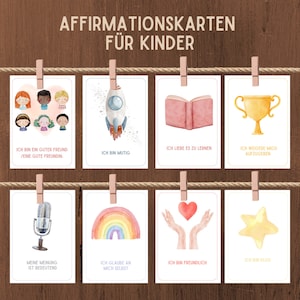 K&ouml;nnte beinhalten: Acht Affirmationskarten mit Aquarell-Illustrationen und positiven Botschaften auf Deutsch. Die Karten zeigen eine Rakete, ein Buch, eine Troph&auml;e, ein Mikrofon, einen Regenbogen, zwei H&auml;nde mit einem Herzen, einen Stern und eine Gruppe von Kindern. Die Karten sind an einer Schnur mit W&auml;scheklammern aufgeh&auml;ngt.