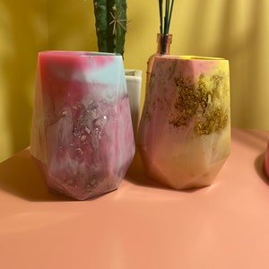 Puede incluir: Dos jarrones decorativos de resina con forma geométrica. Uno es rosa, blanco y azul con motas doradas, el otro amarillo, rosa y blanco con motas doradas. Los jarrones están sobre una superficie rosa.