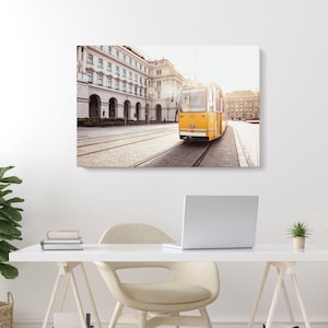 Puede incluir: Un tranvía amarillo vintage viaja por una calle de la ciudad con un gran edificio en el fondo. El tranvía está en una vía y el sol brilla intensamente.