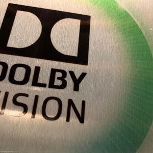 Puede incluir: Una superficie de metal plateado con un logotipo Dolby Vision negro. El logotipo es una "D" estilizada dentro de un cuadrado con las palabras "Dolby Vision" debajo.