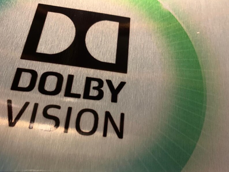 Puede incluir: Una superficie de metal plateado con un logotipo Dolby Vision negro. El logotipo es una "D" estilizada dentro de un cuadrado con las palabras "Dolby Vision" debajo.