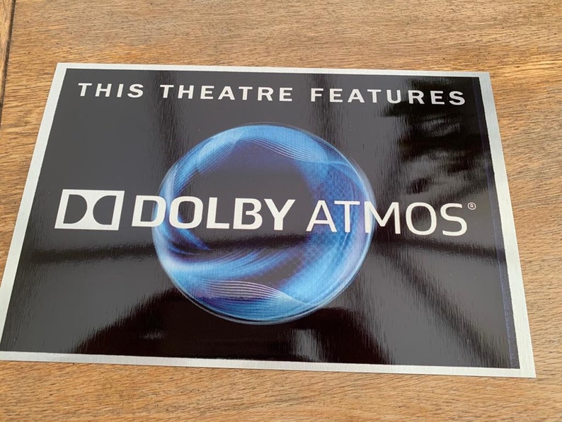 May include: A silver sign with a black background and white text that reads "This theatre features Dolby Atmos". A blue and black sphere with a white outline is in the centre of the sign.