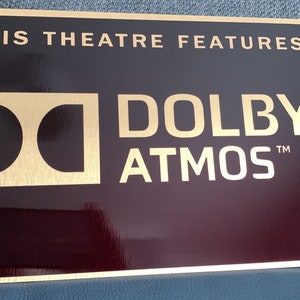 May include: A gold plaque with a black background that reads "This theatre features Dolby Atmos". The Dolby logo is a stylised "D" in a square.