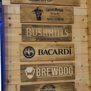 May include: Six wooden signs with various brand names and logos. The signs are arranged vertically on a wood panel wall. The brands include Gordon's London Dry Gin, Captain Morgan Original Spiced Rum, Bushmills Whiskey Distillery, Bacardi, Brewdog Craft Beer for the People, and Peroni Nastro Azzurro.