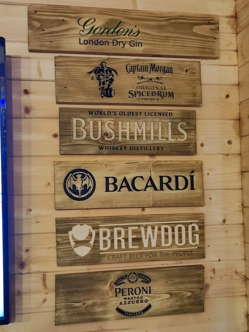 May include: Six wooden signs with various brand names and logos. The signs are arranged vertically on a wood panel wall. The brands include Gordon's London Dry Gin, Captain Morgan Original Spiced Rum, Bushmills Whiskey Distillery, Bacardi, Brewdog Craft Beer for the People, and Peroni Nastro Azzurro.
