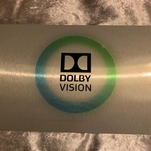Puede incluir: Una placa de metal plateado con un logotipo de Dolby Vision negro en un c&iacute;rculo con un degradado azul y verde. El logotipo est&aacute; rodeado por un halo blanco.