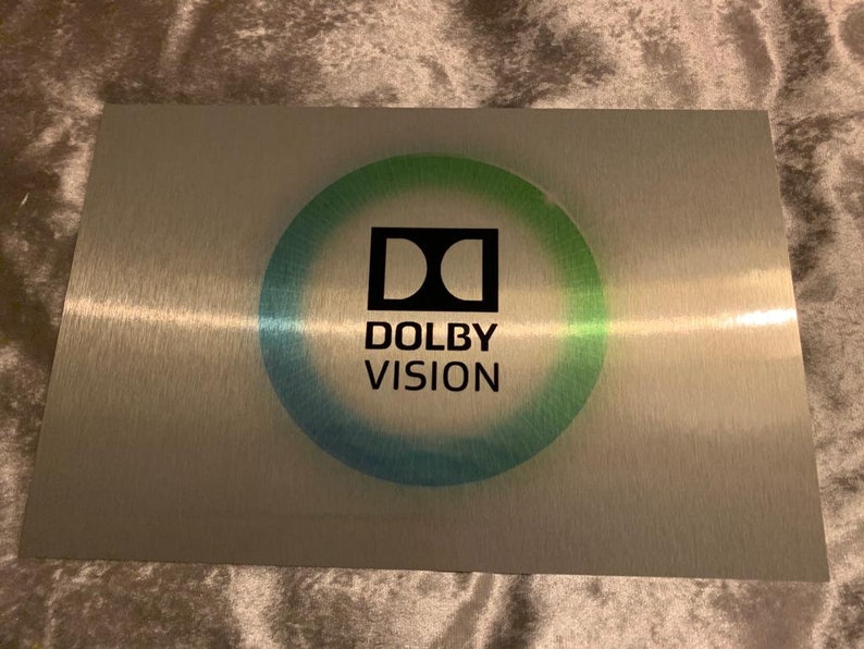 Puede incluir: Una placa de metal plateado con un logotipo de Dolby Vision negro en un c&iacute;rculo con un degradado azul y verde. El logotipo est&aacute; rodeado por un halo blanco.