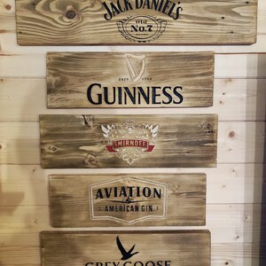 May include: Five wooden signs with different alcohol brand logos. The top sign is brown with black text that reads "Jack Daniels Old No.7 Brand". The second sign is brown with black text that reads "Est. 1759 Guinness". The third sign is brown with a red and white logo that reads "Smirnoff". The fourth sign is brown with white text that reads "Aviation American Gin". The bottom sign is brown with black text that reads "Grey Goose Vodka".