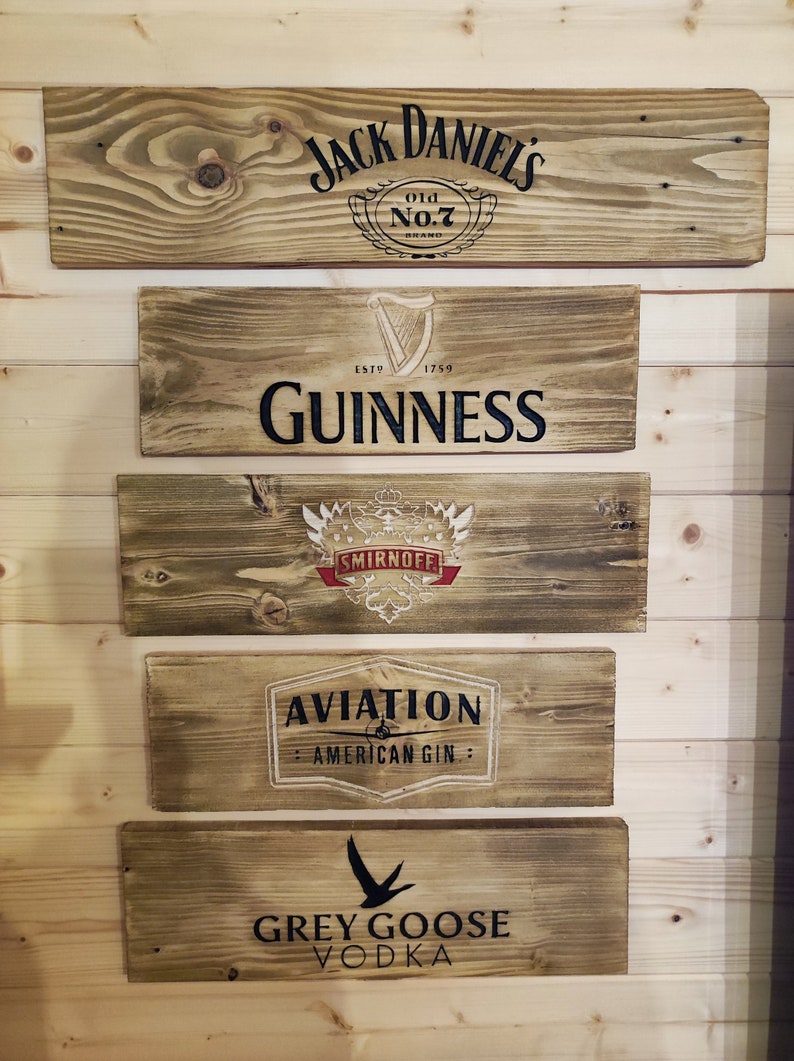 May include: Five wooden signs with different alcohol brand logos. The top sign is brown with black text that reads "Jack Daniels Old No.7 Brand". The second sign is brown with black text that reads "Est. 1759 Guinness". The third sign is brown with a red and white logo that reads "Smirnoff". The fourth sign is brown with white text that reads "Aviation American Gin". The bottom sign is brown with black text that reads "Grey Goose Vodka".