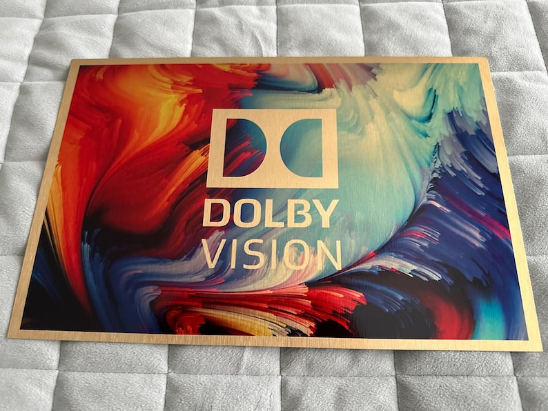 Puede incluir: Cartel rectangular de madera con borde dorado. El cartel presenta el logotipo de Dolby Vision en dorado, con un fondo abstracto colorido de remolinos rojos, azules y turquesas. El texto "DOLBY VISION" tambi&eacute;n es dorado.