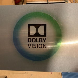 Puede incluir: Una placa de metal plateado con un logotipo de Dolby Vision negro en un c&iacute;rculo verde y azul.