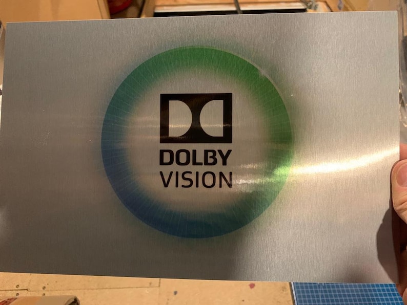 Puede incluir: Una placa de metal plateado con un logotipo de Dolby Vision negro en un c&iacute;rculo verde y azul.
