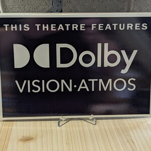 May include: A rectangular sign with a black background and white text. The sign reads "THIS THEATRE FEATURES" at the top, with "Dolby VISION&bull;ATMOS" in a bold, stylized font. The sign is displayed on a clear acrylic stand.