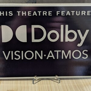 May include: A rectangular sign with the text "THIS THEATRE FEATURES" above the Dolby logo and "VISION.ATMOS" in white lettering. The sign has a dark background and a silver frame, displayed on a wooden surface.