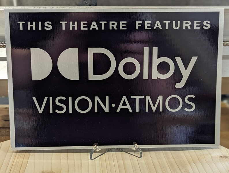 May include: A rectangular sign with the text "THIS THEATRE FEATURES" above the Dolby logo and "VISION.ATMOS" in white lettering. The sign has a dark background and a silver frame, displayed on a wooden surface.