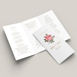 以下が含まれることがあります： マリア＆サイモンの名前とピンクのバラのイラストが描かれた結婚式プログラム。プログラムには、結婚式の日付である2023年8月23日と「ガーデンセレモニー」の文字が記載されています。