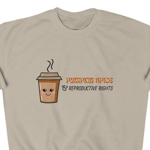 May include: A tan sweatshirt with the text "Pumpkin Spice & Reproductive Rights" in orange. A cartoon coffee cup with a smiling face is on the left. The sweatshirt is a neutral color and has a crew neck.