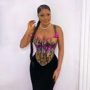 May include: A woman wearing a black skirt and a colorful corset with pink accents. The corset has a tie at the top and a tie at the bottom. She is wearing a gold chain necklace.