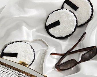 May include: Three white and black reusable makeup remover pads on a white satin surface. A pair of brown sunglasses and a white and gold purse are also visible.
