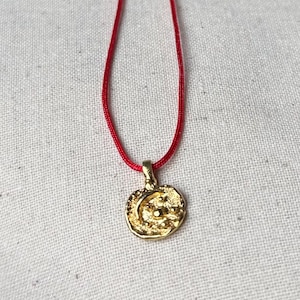 Può includere: Una collana con pendente dorato e cordino rosso. Il pendente rotondo presenta una superficie strutturata e un design a mezzaluna e stelle. La collana è presentata su uno sfondo di tessuto bianco.
