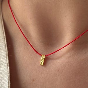 Puede incluir: Un collar con colgante rectangular dorado sobre un cordón rojo. El colgante tiene una superficie texturizada y está suspendido de un fino cordón rojo brillante. El collar se lleva sobre un fondo de tono neutro.