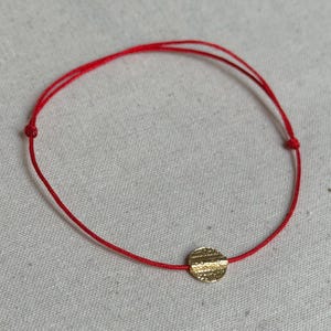 Pode incluir: Uma pulseira fina de cordão vermelho com um pequeno pendente circular de tom dourado e superfície texturizada. A pulseira inclui dois nós deslizantes ajustáveis em cada lado do pendente central para permitir um ajuste personalizado.