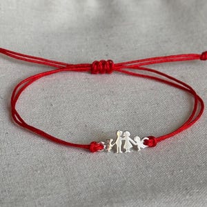 May include: A red cord bracelet featuring a silver-toned charm depicting a family of four figures holding hands. The bracelet includes an adjustable sliding knot closure mechanism, allowing the cord to be tightened or loosened for a secure fit.