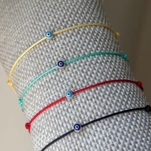 May include: A collection of five colorful string bracelets, each featuring a small, round evil eye charm. The bracelets are in yellow, teal, red, and black. The evil eye charms have a blue center and silver-colored metal around them.