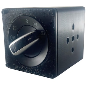 Puede incluir: Un interruptor de luces de coche negro, con forma de cubo, con un dial redondo y una palanca plateada. El dial tiene marcas para diferentes configuraciones de luces, incluyendo faros y luces antiniebla. El cubo tiene varios agujeros pequeños en un lado.