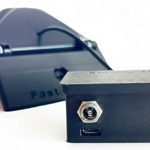 May include: Black electronic device with a rectangular box shape and a smaller, angled component behind it. The box has a port and a USB port. The angled component has the word "Fast" printed on it.