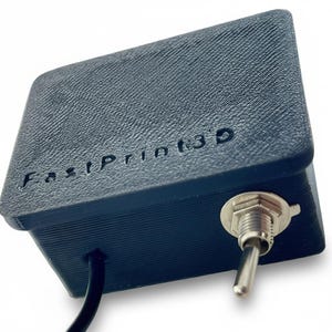 May include: A dark blue, rectangular 3D-printed device with the text "FastPrint3D" embossed on the top. It features a silver toggle switch on the side and a black cable extending from the bottom. The device has a textured surface.