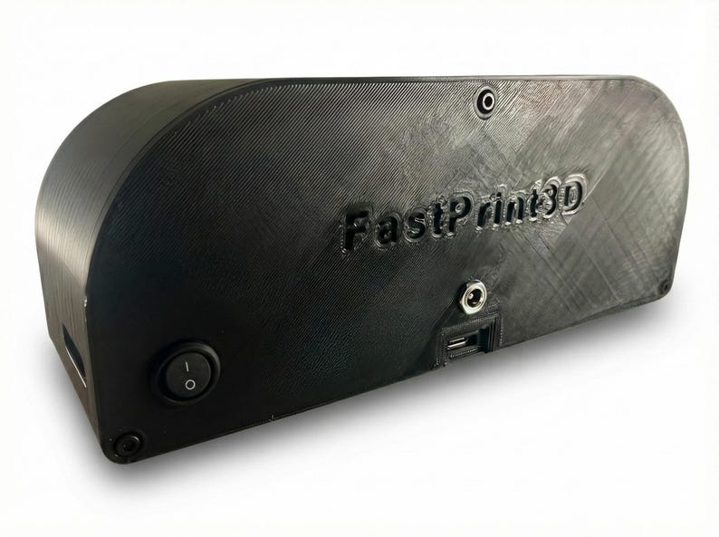 May include: Black 3D printer enclosure with the text "FastPrint3D" embossed on the side. The enclosure has a rounded top and features a power button, a USB port, and other ports. The device is designed to house a 3D printer.