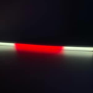 May include: A long, rectangular light bar with a gradient of colors. One end is white, transitioning to red in the center, and back to white. The bar has black end caps and is illuminated against a dark background.
