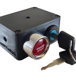 Op de afbeelding: Een zwart rechthoekig apparaat met een rode "ENGINE START" knop en een chromen rand. Een sleutel is in een sleutelgat gestoken met de tekst "OFF" en "ON". Het apparaat heeft de tekst "FastPrint3D" aan de zijkant.