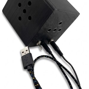 K&ouml;nnte beinhalten: Ein schwarzes, w&uuml;rfelf&ouml;rmiges elektronisches Ger&auml;t mit mehreren kreisf&ouml;rmigen &Ouml;ffnungen. Es verf&uuml;gt &uuml;ber einen Kippschalter, mehrere Eingangsanschl&uuml;sse und ein USB-Kabel mit einem gemusterten Kabel. Das Ger&auml;t scheint eine kundenspezifische elektronische Komponente oder ein Gadget zu sein.