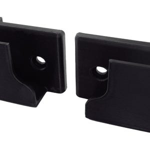 Könnte beinhalten: Zwei schwarze, rechteckige Kunststoffhalterungen. Jede Halterung hat eine Aussparung und ein kreisförmiges Loch. Die Halterungen sind so konzipiert, dass sie einen Gegenstand halten und an einer Oberfläche befestigt werden können.