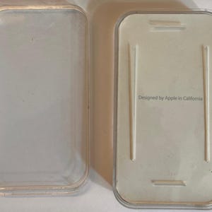 May include: A clear, rectangular plastic case, open, with a white interior. The case is designed to hold a small electronic device. The text "Designed by Apple in California" is printed on the interior.