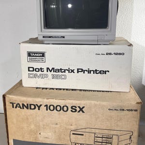 Könnte beinhalten: Ein Vintage-Tandy-Computer-Setup mit einem grauen Monitor auf Kartons für einen Dot Matrix Printer DMP 130 und einen Tandy 1000 SX Personal Computer. Die Kartons zeigen Produktnamen und Modellnummern.