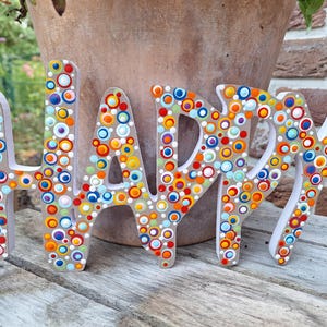 Könnte beinhalten: Ein Holzschild mit dem Wort "HAPPY" in Großbuchstaben. Jeder Buchstabe ist weiß bemalt und mit bunten, gepunkteten Kreisen in Rot-, Orange-, Gelb-, Blau- und Grüntönen verziert. Das Schild steht auf einer Holzoberfläche.