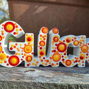Könnte beinhalten: Ein weißes Holzschild mit dem Wort "Glück" in orange, gelben und roten Punktmustern bemalt. Das Schild steht auf einer verwitterten Holzoberfläche, mit einem verschwommenen Hintergrund aus einem Terrakottatopf und Grün.